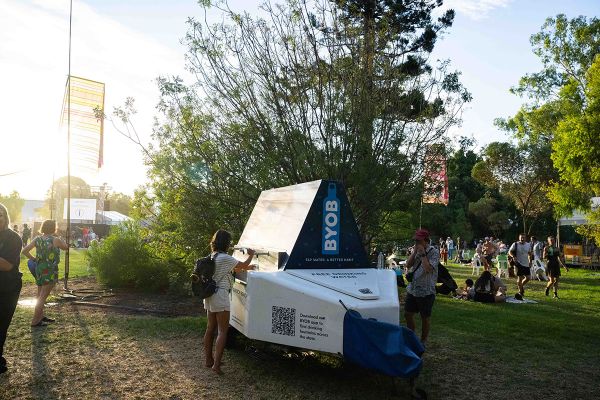 People using a SA Water Quench bench at WOMADelaide — a woman with a backpack fills her water bottle on one side, a man fills his bottle on the other, with event goers in the background and afternoon sunlight shining through the trees.
